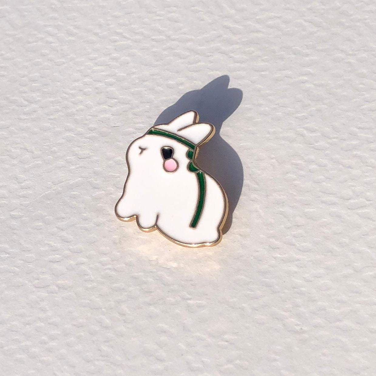 Ghost-shaped enamel pin with a green headband on a white textured background
