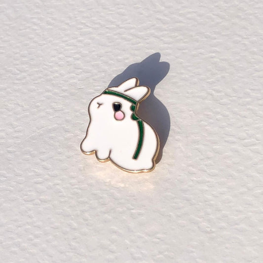 Ghost-shaped enamel pin with a green headband on a white textured background